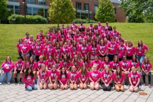 A large group of students and teachers are sitting on a ledge and in the grass on a hill. They are all wearing matching pink T-shirts. Photo from Media Now JAM 2022 at Webster University in St. Louis