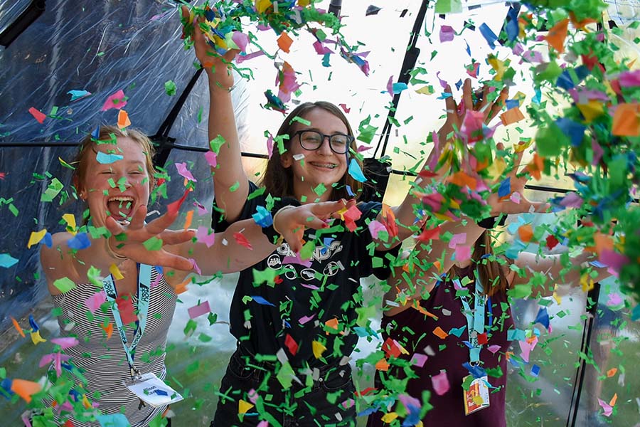 Three smiling students in a tent throwing confetti in the air